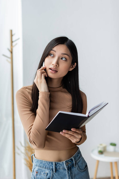 азіатська жінка з копією книги розмовляє на смартфоні і дивиться вдома
 - Фото, зображення