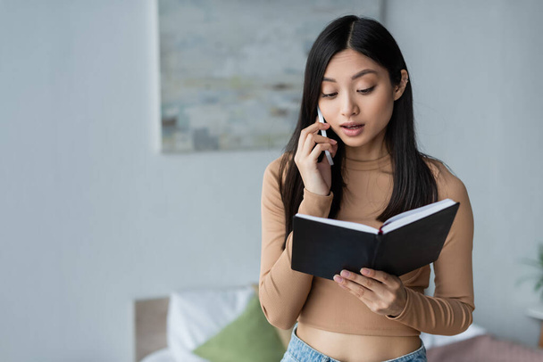 young asian woman looking in notebook while talking on cellphone in bedroom - Foto, Imagem