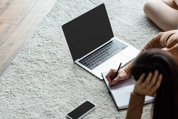 overhead view of blurred woman writing in notebook near laptop and smartphone on floor carpet - Photo, Image