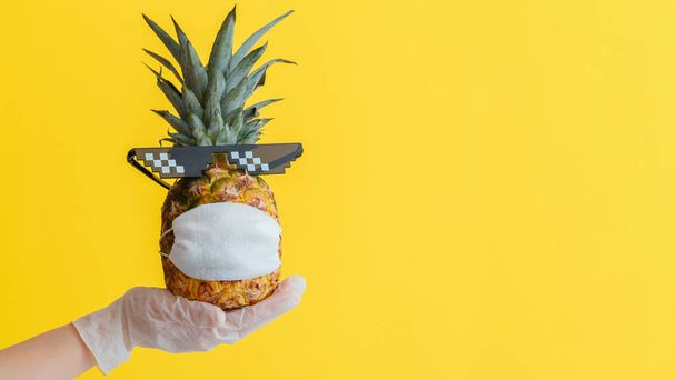 Funny pineapple face in a protective medical mask. Doctor hand in rubber glove holds summer tropical fruit pineapple in sunglasses. Safe travel concept during covid coronavirus lockdown. Long web - Foto, imagen