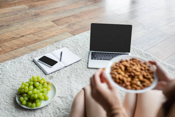 partial view of blurred freelancer holding bowl with almonds near gadgets and fresh grape on floor - Foto, Imagen