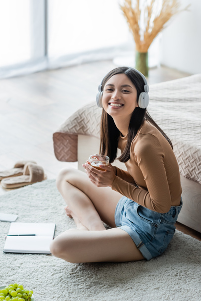 cheerful asian woman in headphones holding cup of tea while sitting on floor with crossed legs near notebook - Photo, Image