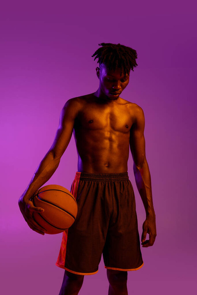 Młody Afrykanin, koszykarz pozujący z piłką na fioletowym neonowym tle. - Zdjęcie, obraz