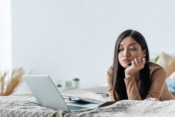 pensive asian woman looking at laptop while lying in bedroom - Photo, image