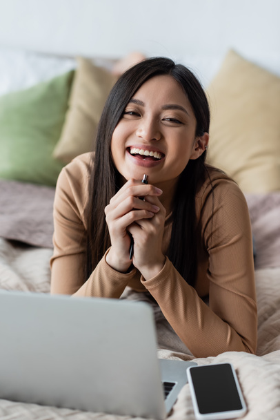 asian woman smiling at camera near blurred smartphone and laptop on bed - Φωτογραφία, εικόνα