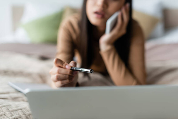blurred woman pointing with pen at laptop while talking on cellphone in bedroom - Photo, image