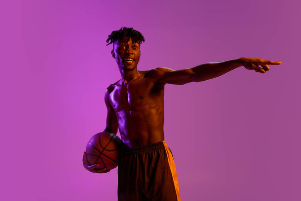 Young african man, basketball player posing with ball isolated over purple neon background. - Фото, зображення