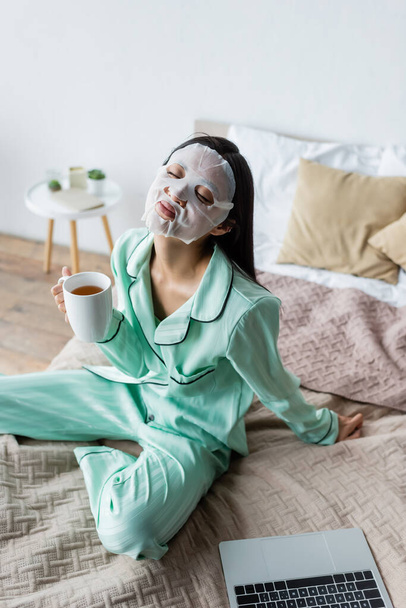 asiatique femme en feuille masque assis sur lit avec tasse de thé et les yeux fermés près ordinateur portable - Photo, image