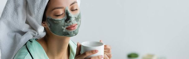 elégedett ázsiai nő agyag maszk az arcon és törölköző a fejen tartó csésze tea, banner - Fotó, kép