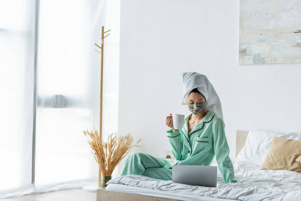 asian woman in clay mask and pajamas sitting on bed with cup of tea and looking at laptop - Foto, Bild