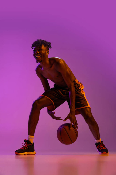 Młody Afrykanin, koszykarz w akcji, ruch z kulą odizolowaną od fioletowego neonowego tła. - Zdjęcie, obraz
