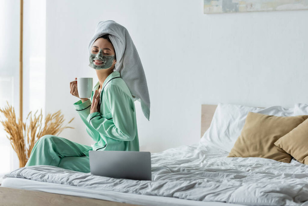 pleased asian woman with closed eyes holding cup of tea while sitting near laptop in bedroom - Foto, Imagen