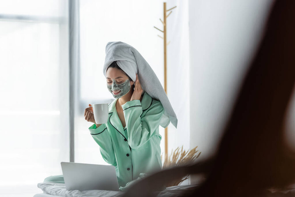 usmívající se asijská žena v pyžamu a hliněné masky sedí s šálkem čaje v blízkosti notebooku v ložnici - Fotografie, Obrázek