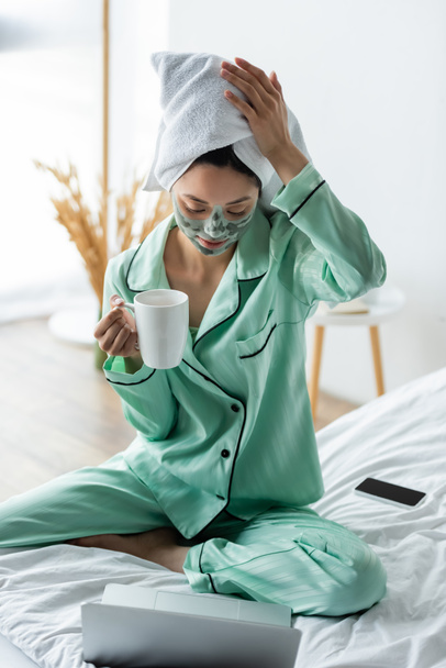 азиатка в глиняной маске и полотенце на голове сидит на кровати с чашкой чая возле ноутбука - Фото, изображение