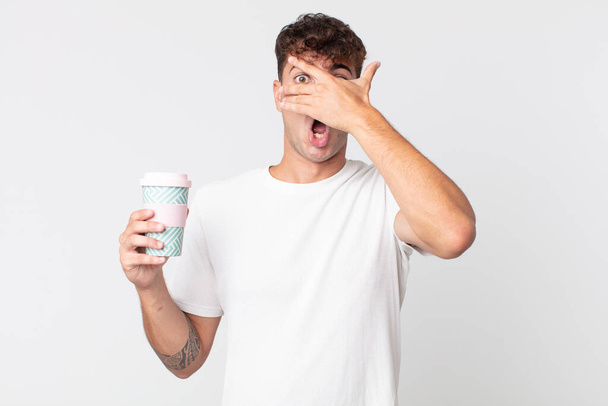 young handsome man looking shocked, scared or terrified, covering face with hand and holding a take away coffee - Fotoğraf, Görsel