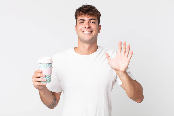 young handsome man smiling happily, waving hand, welcoming and greeting you and holding a take away coffee - Photo, image