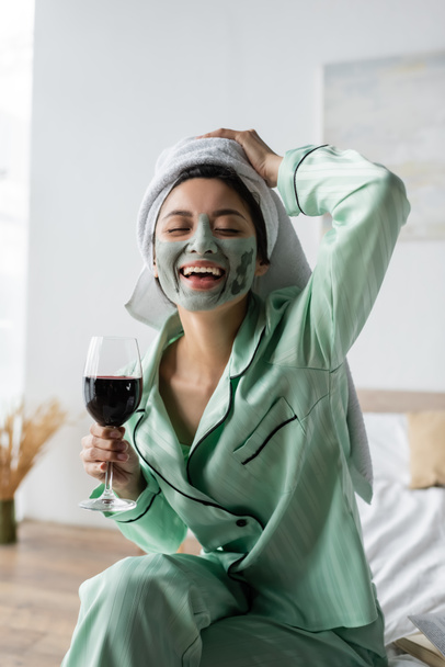excited asian woman in clay mask and towel on head holding glass of red wine and laughing with closed eyes - Photo, Image