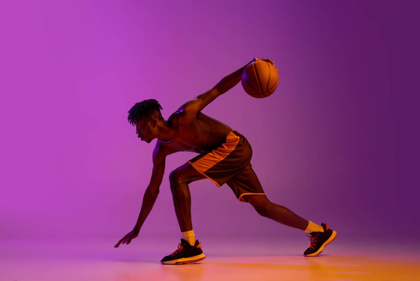 Young african man, basketball player in action, motion with ball isolated over purple neon background. Side view - 写真・画像