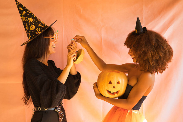 Молоді іспаномовні та латиноамериканські жінки, одягнені як відьми, голосно сміються і роздають цукерки з Хелловін на рожевому фоні.. - Фото, зображення