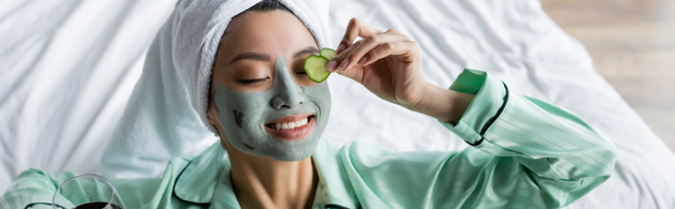 joyful asian woman in clay mask applying fresh cucumber slices on eye, banner - Foto, Imagen