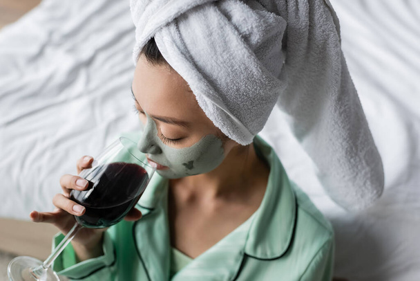 high angle view of asian woman in clay mask and towel on head drinking red wine in bedroom - Fotó, kép