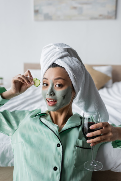 young asian woman in clay mask and pajamas holding cucumber slices and glass of red wine while looking at camera - Valokuva, kuva