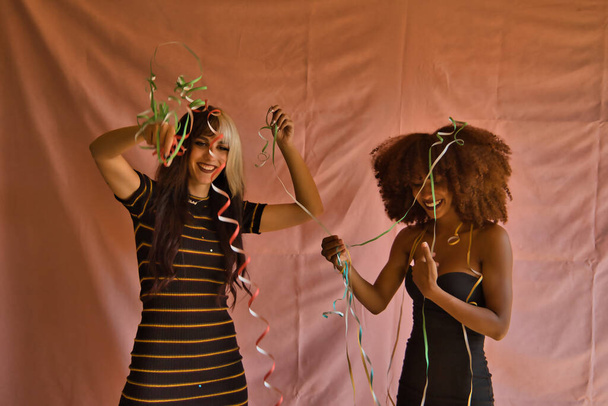 Fiatal spanyol és latin nők portréja egy partin, akik szalaggal dobálják egymást, rózsaszín alapon.. - Fotó, kép