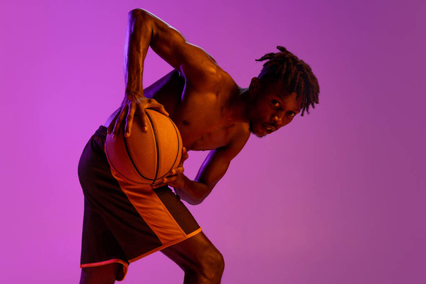Junge afrikanische Mann, Basketballspieler posiert mit Ball isoliert über lila Neon Hintergrund. - Foto, Bild