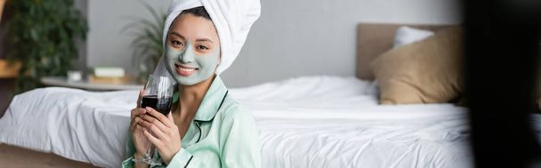 cheerful asian woman with clay mask and towel on head holding glass of red wine in bedroom, banner - Photo, image