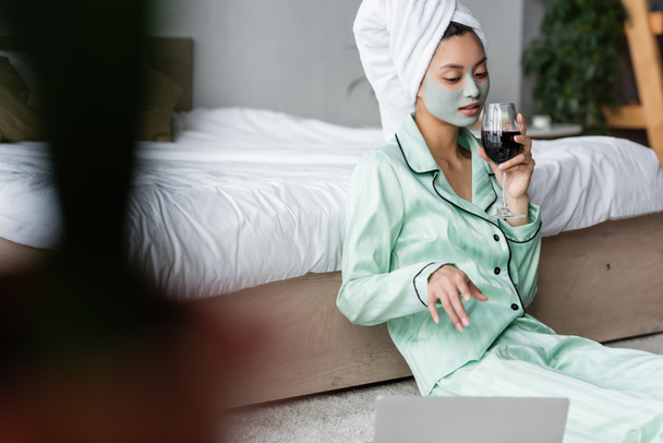 mulher asiática em pijama e máscara de barro sentado no chão e apontando para laptop em primeiro plano borrado - Foto, Imagem