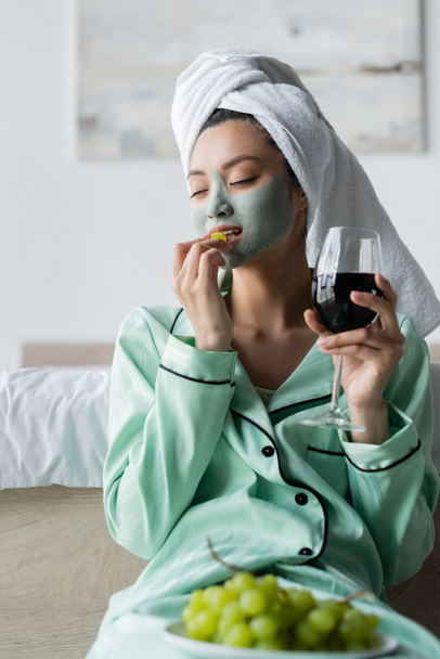 asian woman in towel on head and clay mask eating grape while holding glass of red wine - Valokuva, kuva