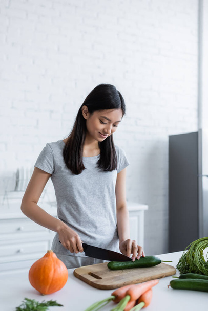positive asian woman cutting fresh cucumber near pumpkin and carrots on kitchen table - Photo, Image