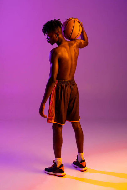 Νεαρός Αφρικανός, μπασκετμπολίστας ποζάρει με μπάλα απομονωμένη πάνω από μωβ νέον φόντο. Πίσω όψη - Φωτογραφία, εικόνα
