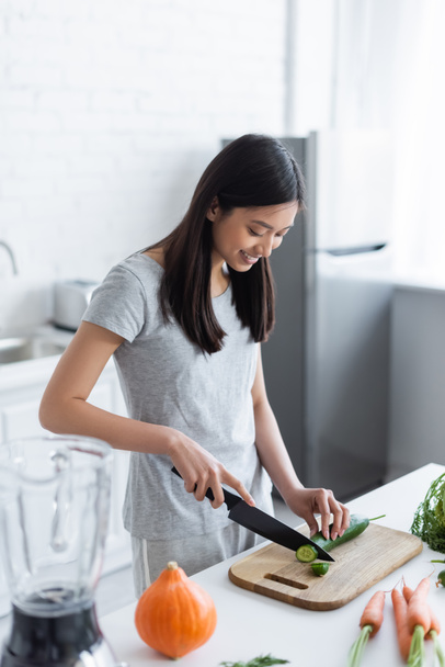 happy asian woman cutting cucumber near fresh carrots and pumpkin in kitchen - Photo, Image