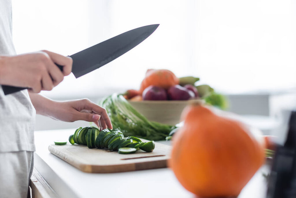 partial view of woman with knife near sliced cucumber on chopping board and pumpkin on blurred foreground - Photo, Image