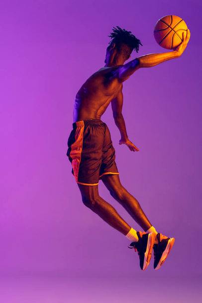 Młody Afrykanin, koszykarz w ruchu z piłką odizolowaną od gradientowego fioletowego neonu tła. - Zdjęcie, obraz