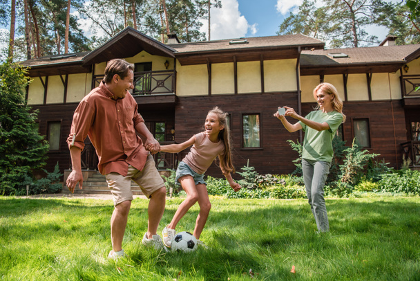 Parent and child playing football near mother with smartphone on lawn  - Foto, immagini
