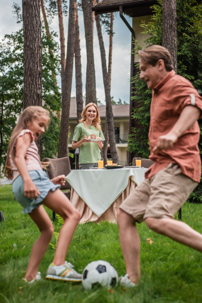 Smiling woman with plate standing near table and blurred family playing football  - Foto, Bild