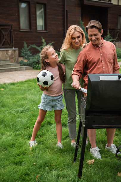 Smiling kid with football looking at parents cooking on grill outdoors  - Zdjęcie, obraz