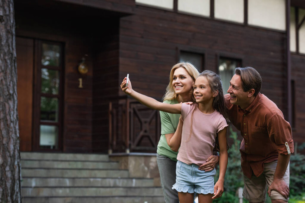 Smiling girl taking selfie with parents near blurred vacation house  - Zdjęcie, obraz
