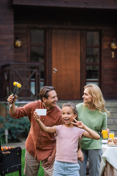 Happy kid showing peace sign while taking selfie near parents with food outdoors  - Foto, Bild