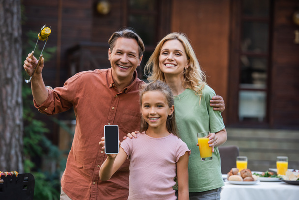 Улыбающийся ребенок показывает смартфон рядом с родителями с едой и апельсиновым соком на открытом воздухе  - Фото, изображение