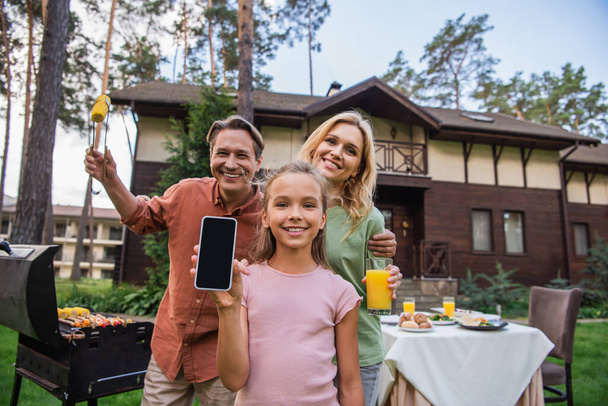 Chica mostrando teléfono inteligente cerca de padres sonrientes con comida y parrilla al aire libre  - Foto, imagen