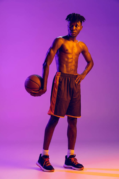 Νεαρός Αφρικανός, μπασκετμπολίστας ποζάρει με μπάλα στο χέρι απομονωμένος σε μωβ φόντο νέον. - Φωτογραφία, εικόνα