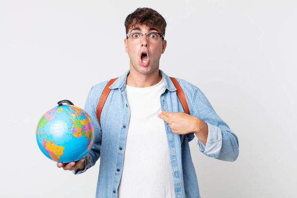young handsome man looking shocked and surprised with mouth wide open, pointing to self. student holding a world globe map - Photo, image