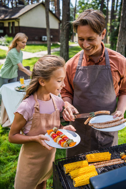 Smiling man holding sausages near daughter with vegetables and grill  - Fotoğraf, Görsel