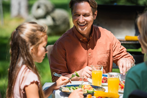 Alegre hombre mirando borrosa hija cerca de la comida y jugo de naranja en la mesa al aire libre  - Foto, imagen