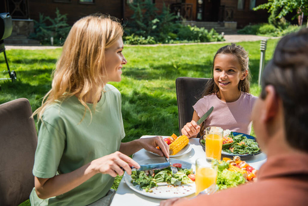 Smiling kid looking at mother with cutlery near food and orange juice outdoors  - Фото, зображення