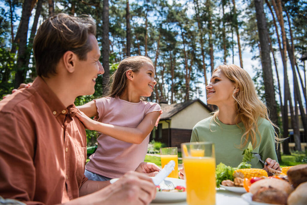 Side view of smiling kid hugging father near mother and tasty food outdoors  - Photo, image