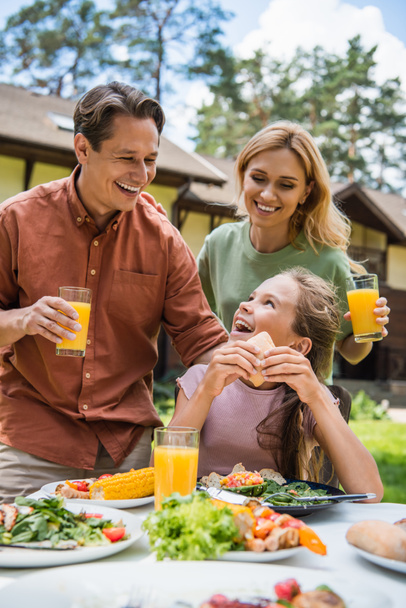 Des parents heureux avec du jus d'orange regardant leur fille près d'une nourriture savoureuse pendant le pique-nique  - Photo, image